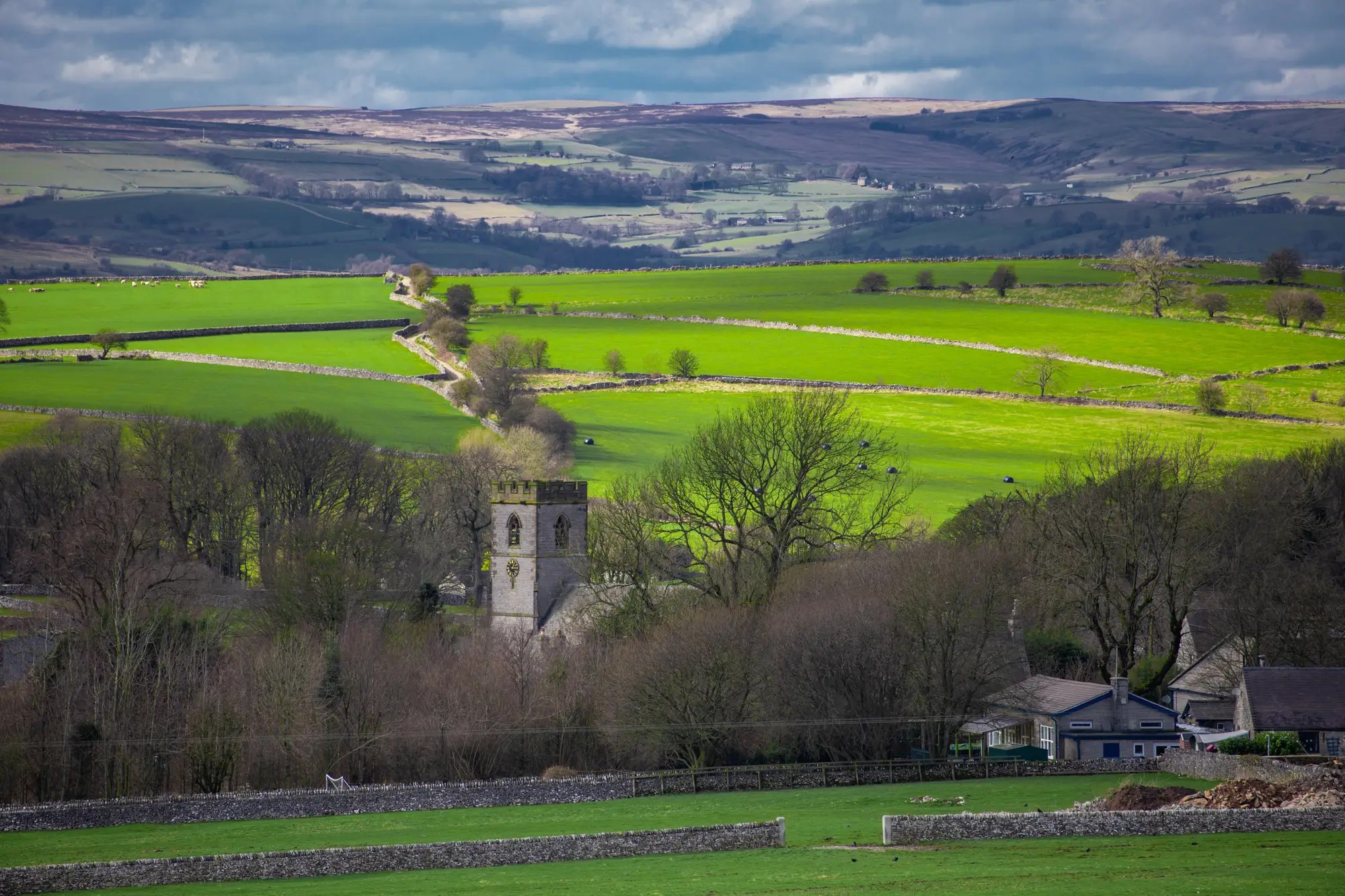 A church tower among bare trees at the edge of a village, with walled pasture fields climbing the valley side behind it